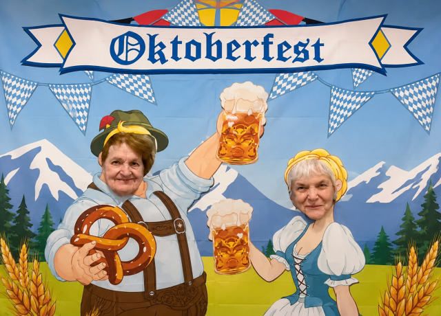 Two Bingley Park residents smiling through an Oktoberfest photo board with beer steins and a pretzel.